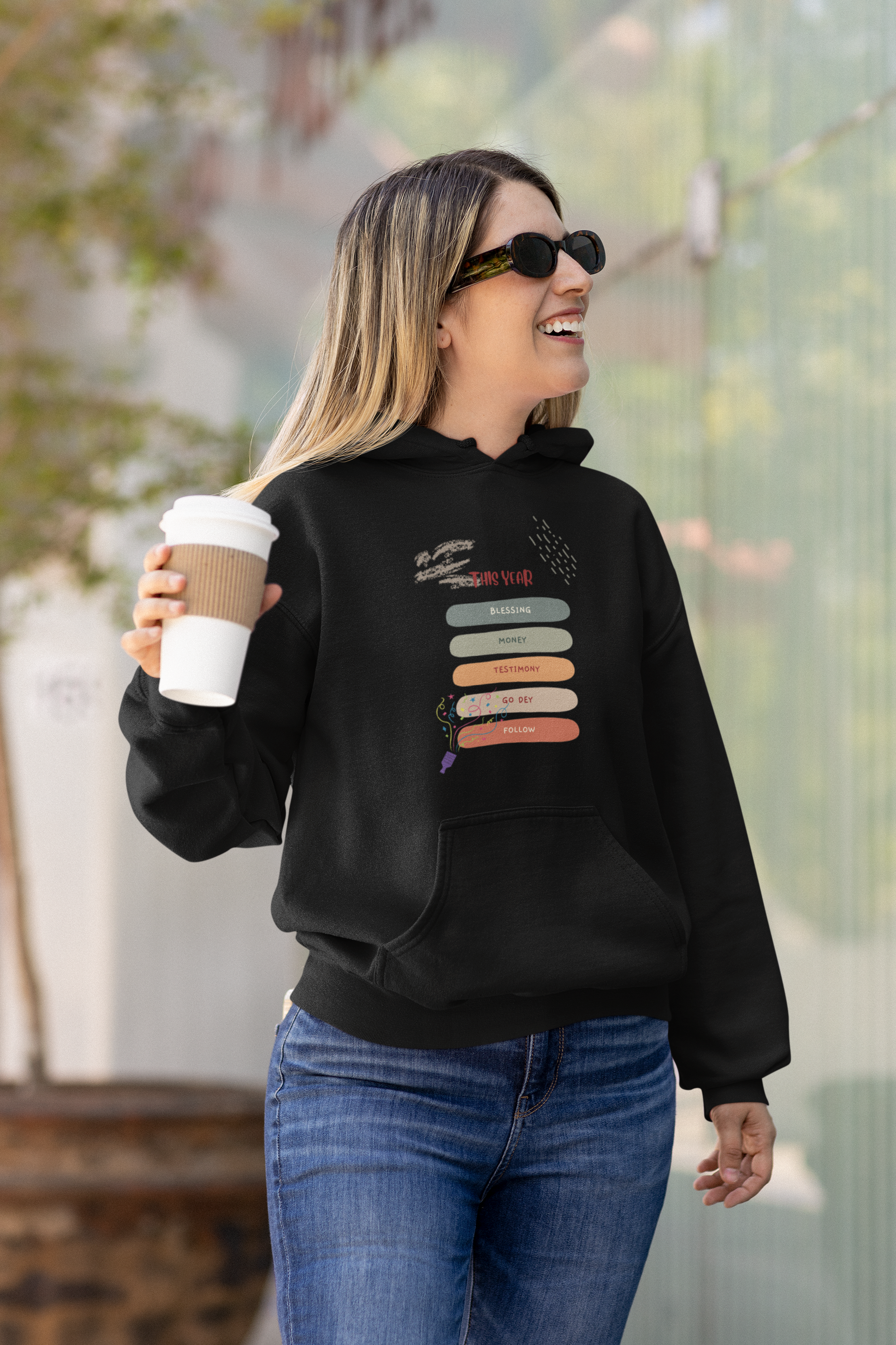 Woman wearing a black hoodie with a colorful design, holding a coffee cup outdoors.