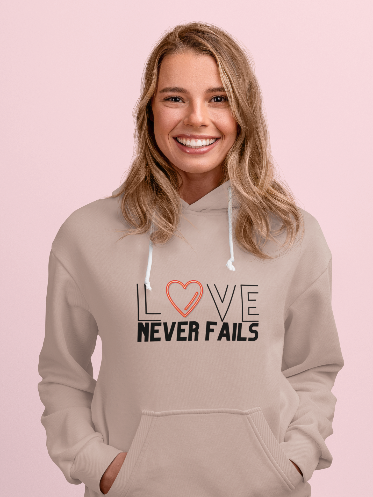 Woman wearing a beige hoodie with 'LOVE NEVER FAILS' text on a pink background
