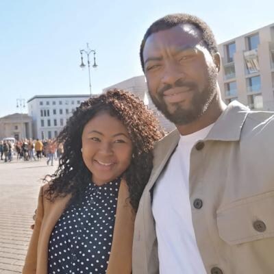 Two people standing together outdoors with a building and people in the background