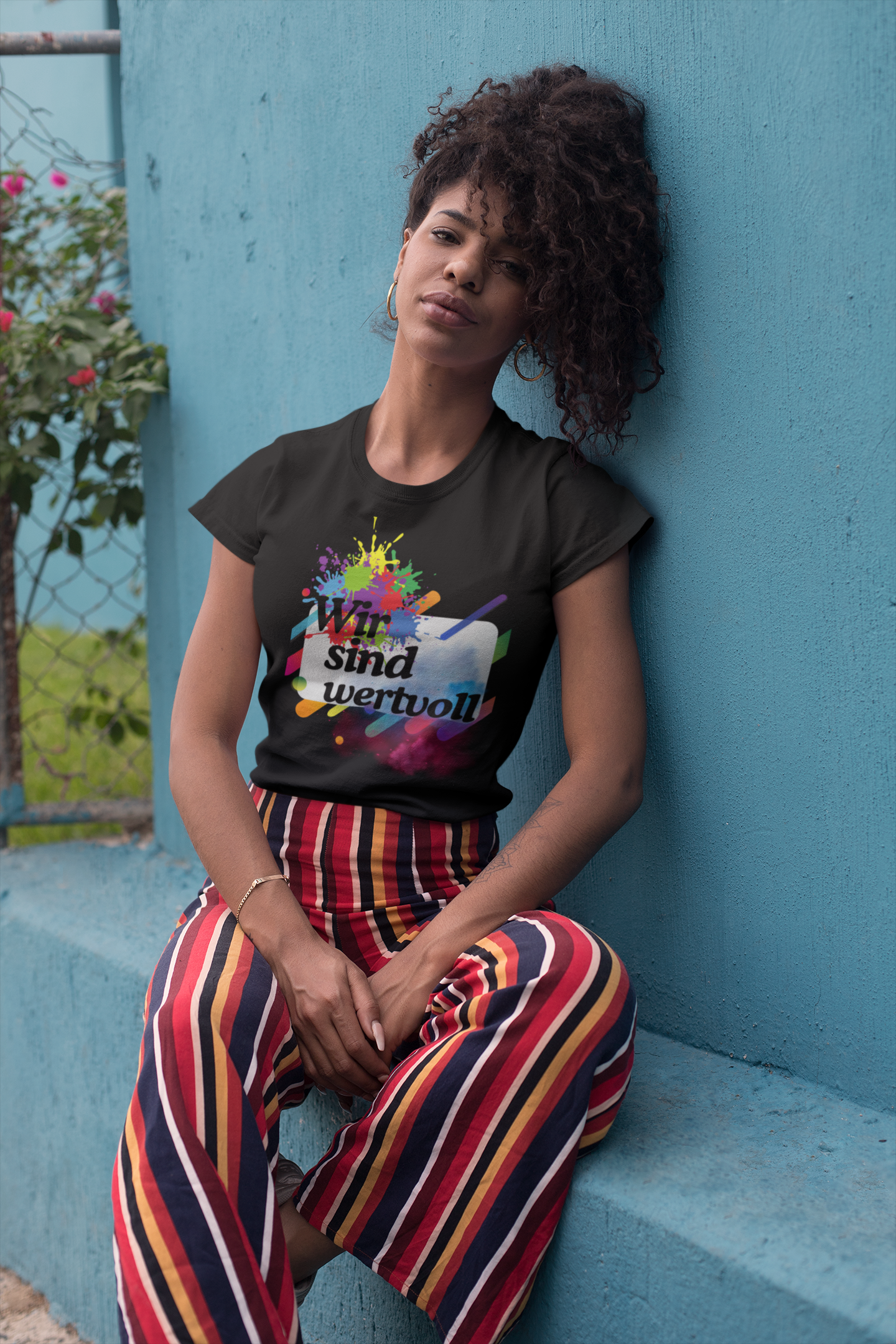Person wearing a black t-shirt with colorful graphics and text, sitting against a blue wall.