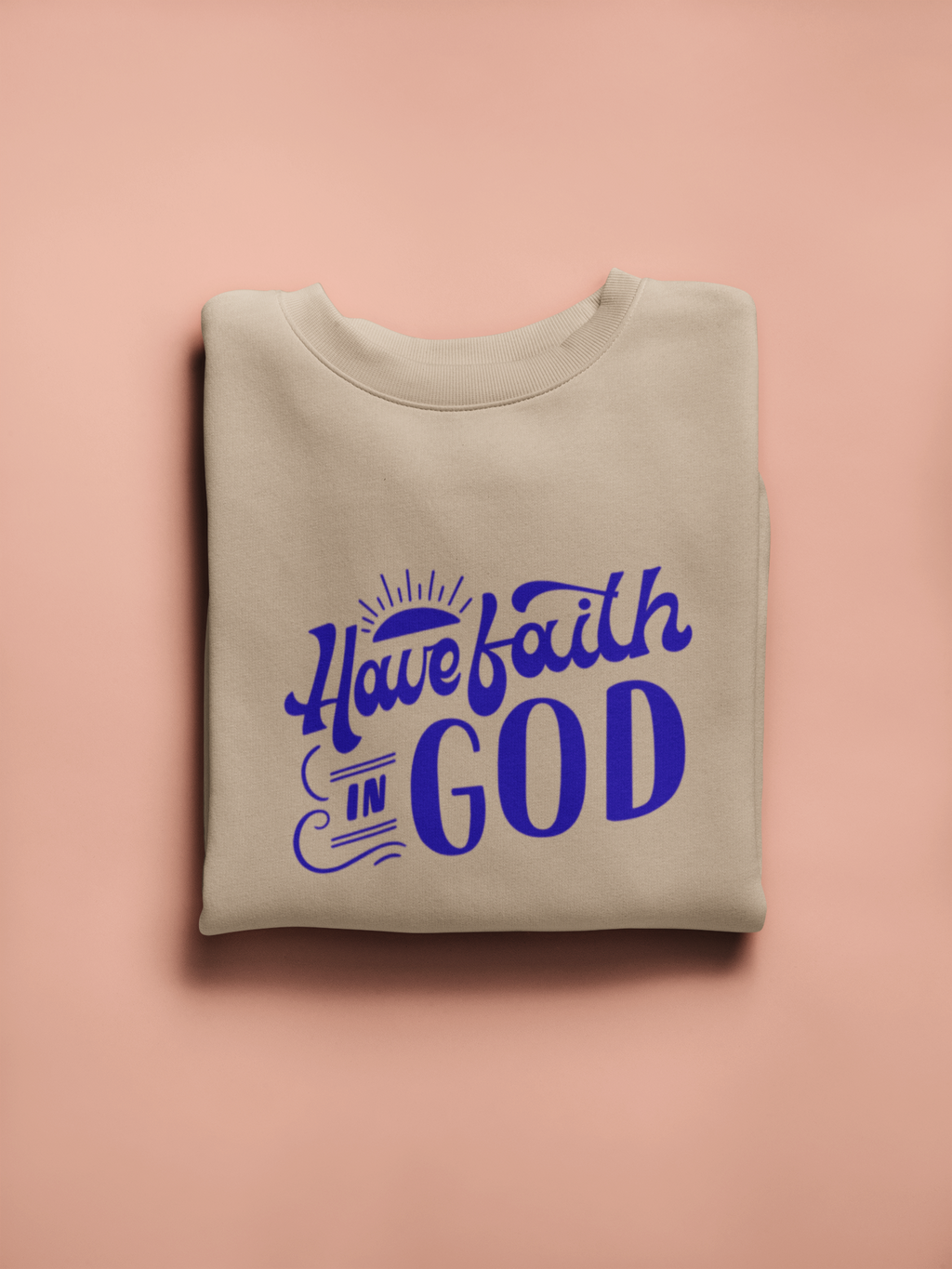 Folded beige sweatshirt with 'Have faith in God' text in purple on a pink background