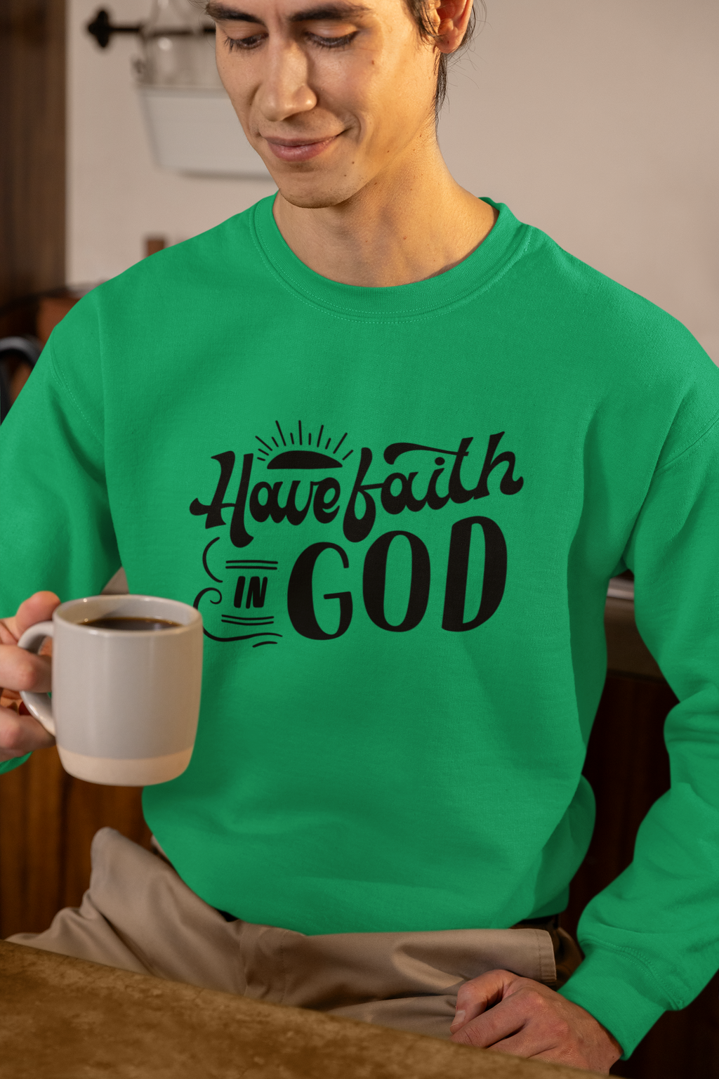 Person wearing a green sweatshirt with 'Have faith in God' text, holding a mug.