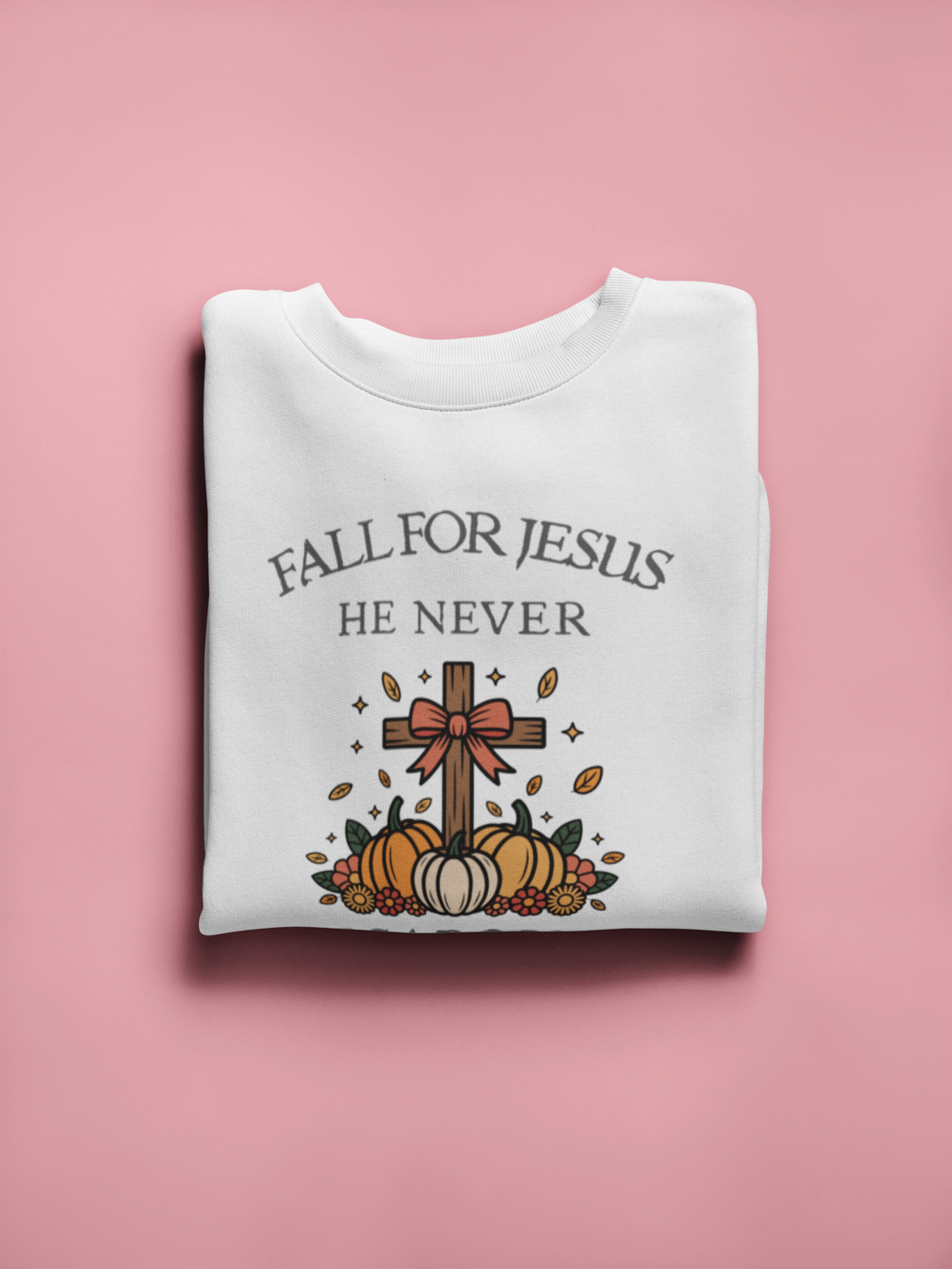 Folded white sweatshirt with 'Fall for Jesus He Never' text and cross design on a pink background