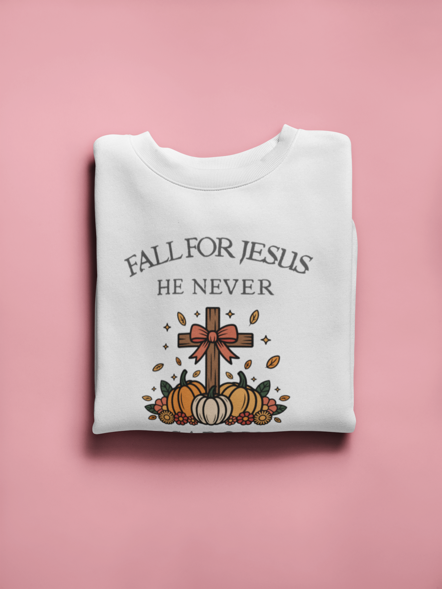 Folded white sweatshirt with 'Fall for Jesus He Never' text and cross design on a pink background
