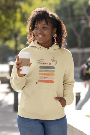 Woman wearing a beige hoodie with colorful text design, holding a coffee cup outdoors.