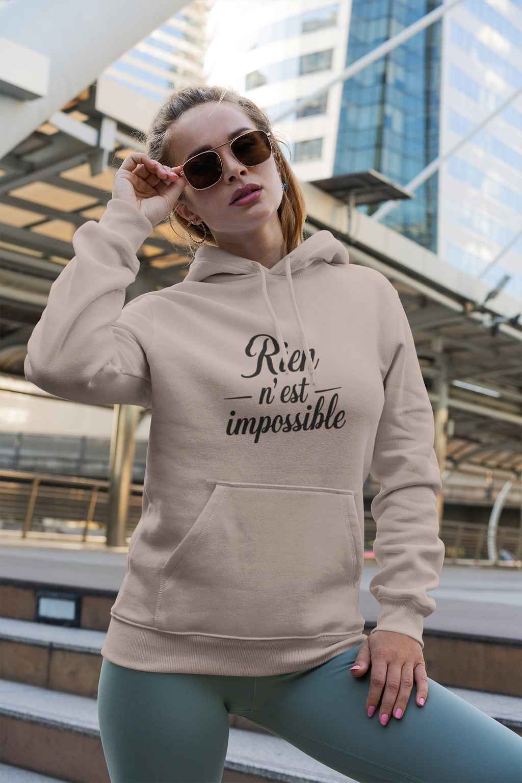 Woman wearing a beige hoodie with text, standing outdoors with modern architecture in the background
