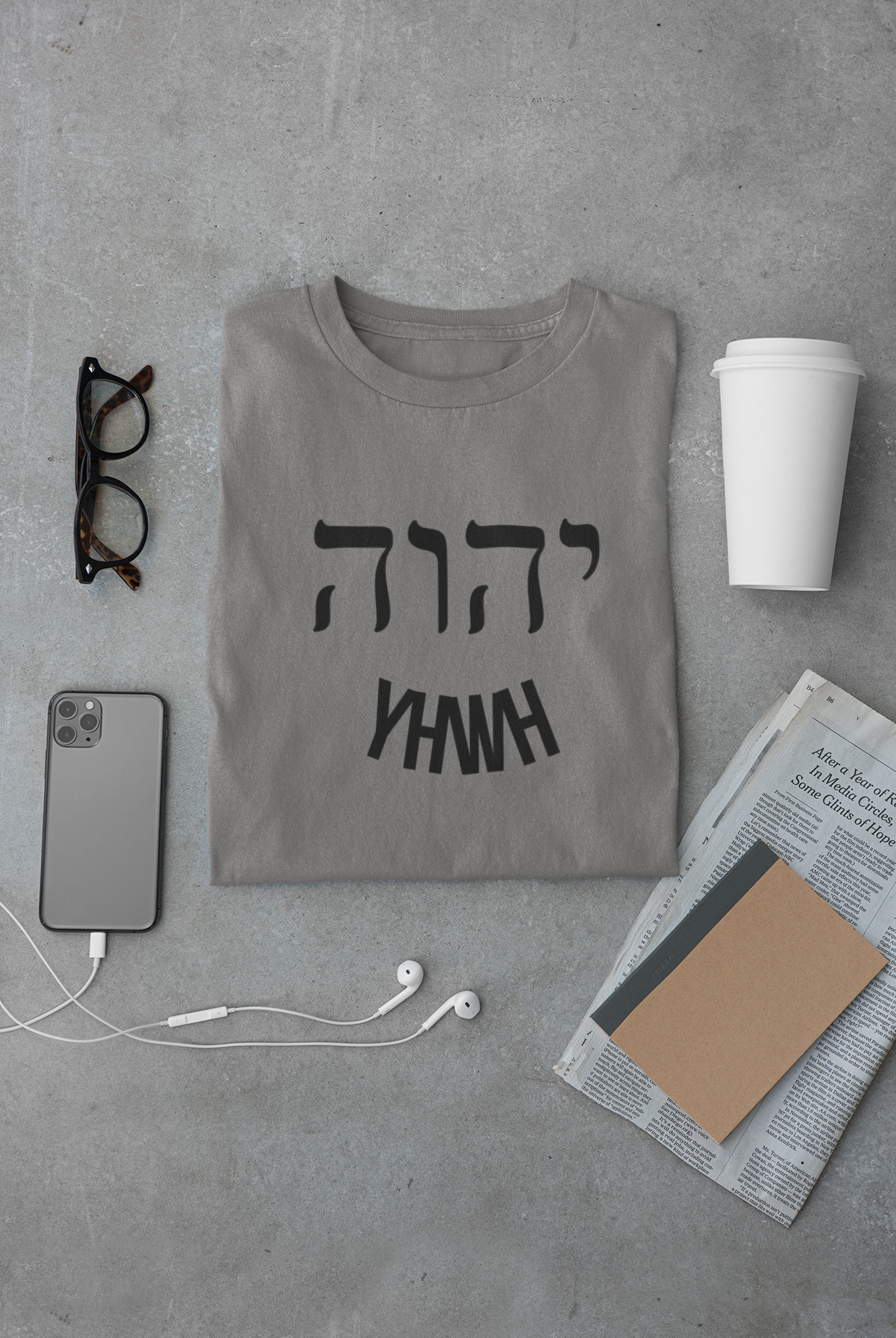 Gray t-shirt with black text on a gray surface with phone, glasses, cup, and newspaper.