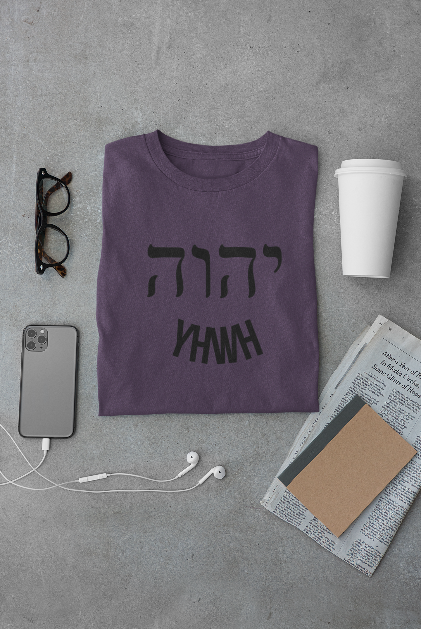 Purple t-shirt with black text on a gray surface with phone, glasses, and newspaper.