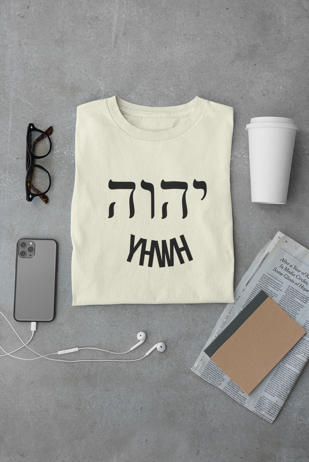 Beige t-shirt with black text on a gray surface with phone, glasses, and cup.