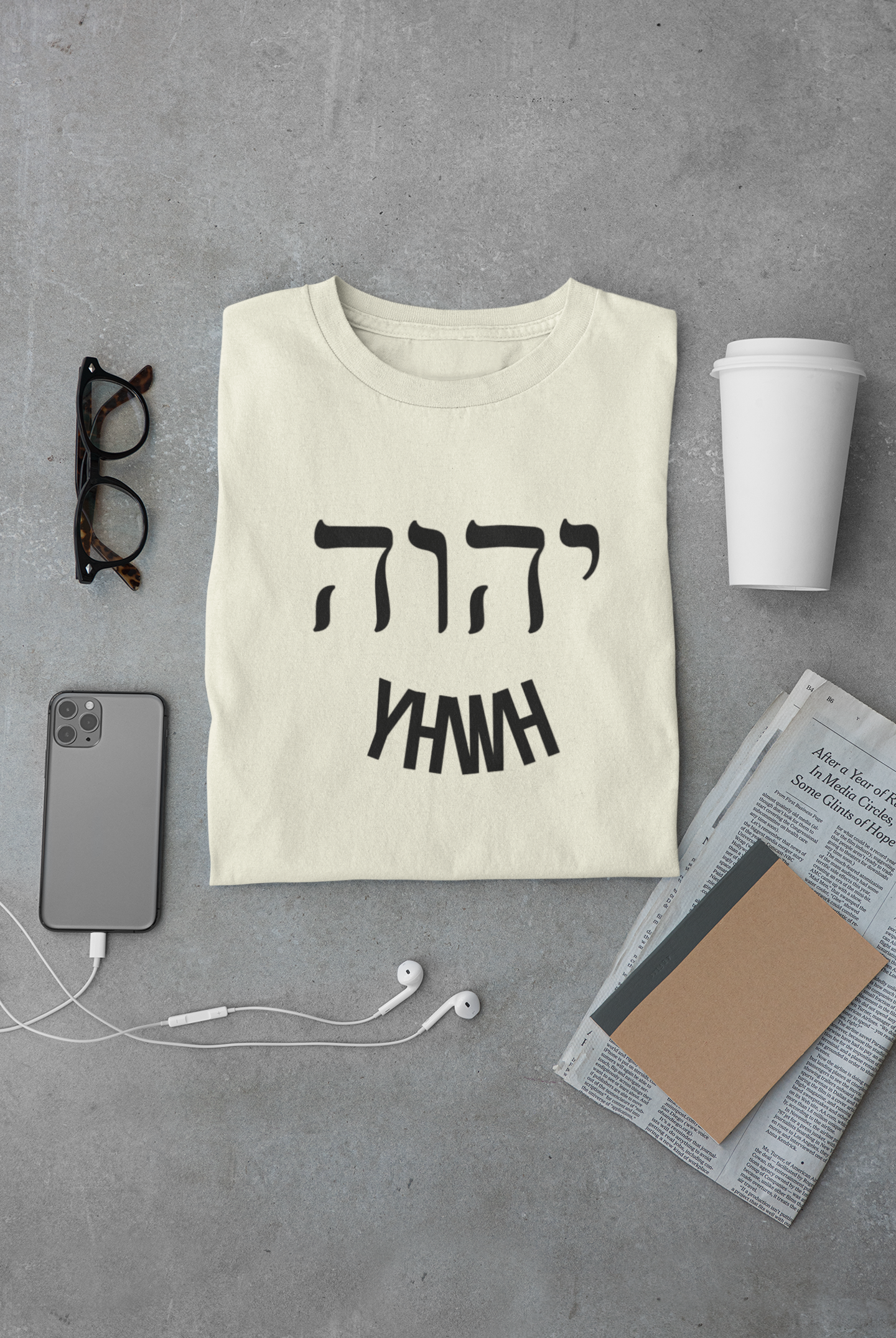 Beige t-shirt with black text on a gray surface with phone, glasses, and cup.