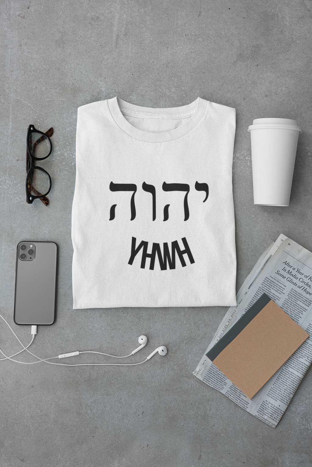 White t-shirt with black text on a gray surface with phone, cup, and glasses.